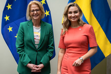 Elisabeth Svantesson och Ebba Busch står sida vid sida och ler mot kameran, framför EU-flaggor och svenska flaggor. Svantesson bär grön kavaj och Busch en korallfärgad klänning.