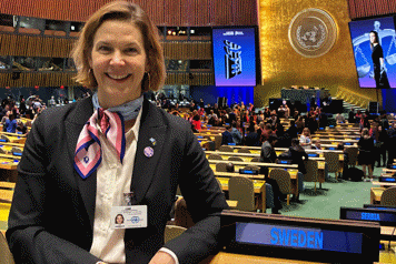 Nina Larsson står i ett FN:s stora mötesrum sidan om en skylt det står SWEDEN på. I bakgrunden syns långa bänkrader och stolar. En stor grupp människor står i mitten av rummet. Väggarna har träpaneler och bakom talarstolen längst bort i mötesrummet är en del av väggen förgylld med FN:s emblem på.