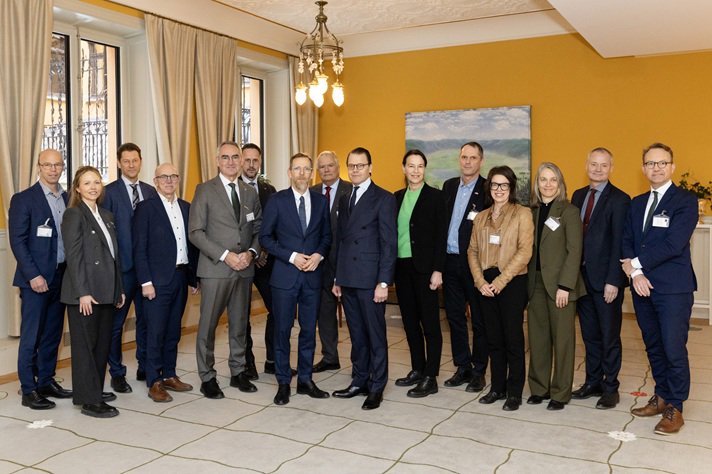 En grupp på femton personer står samlade i ett mötesrum med gula väggar och stora fönster. De är formellt klädda och står uppställda i en halvcirkel för ett gruppfoto.