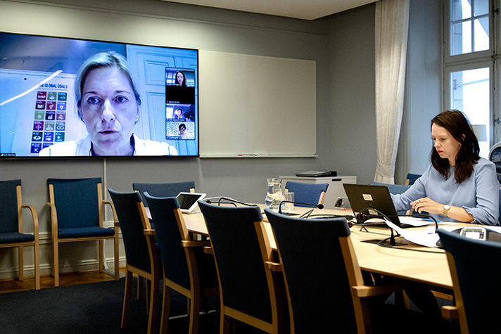 Åsa Lindhagen sitter i ett mötesrum och tittar på sin dator där ett digitalt möte pågår. På en storbildsskärm syns deltagare från mötet.