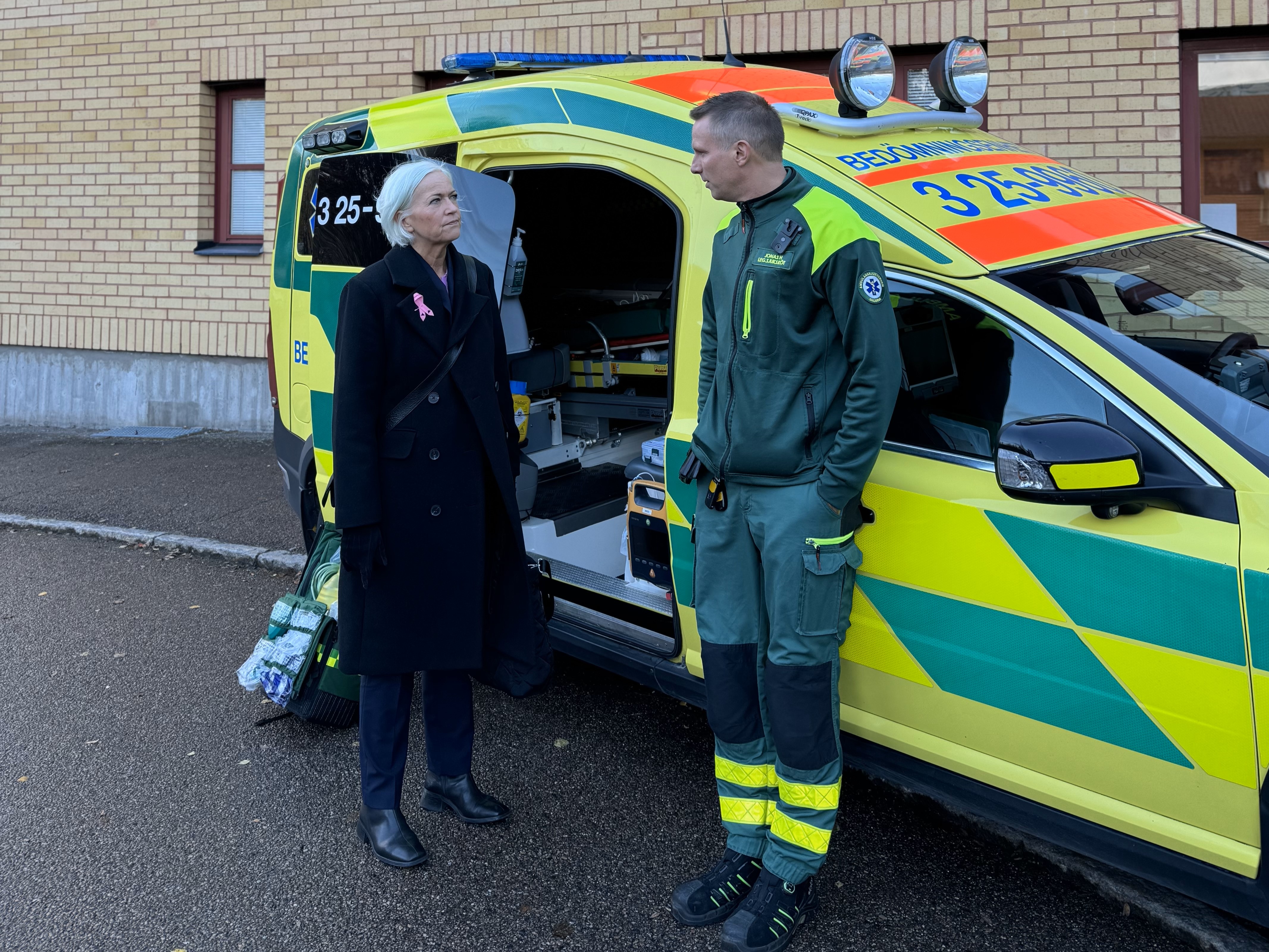 Sjukvårdsminister Acko Ankarberg Johansson står bredvid en ambulans. Bredvid henne, lutad mot ambulansen, står en arbetsklädd man med händerna i fickorna som tittar på henne och pratar.