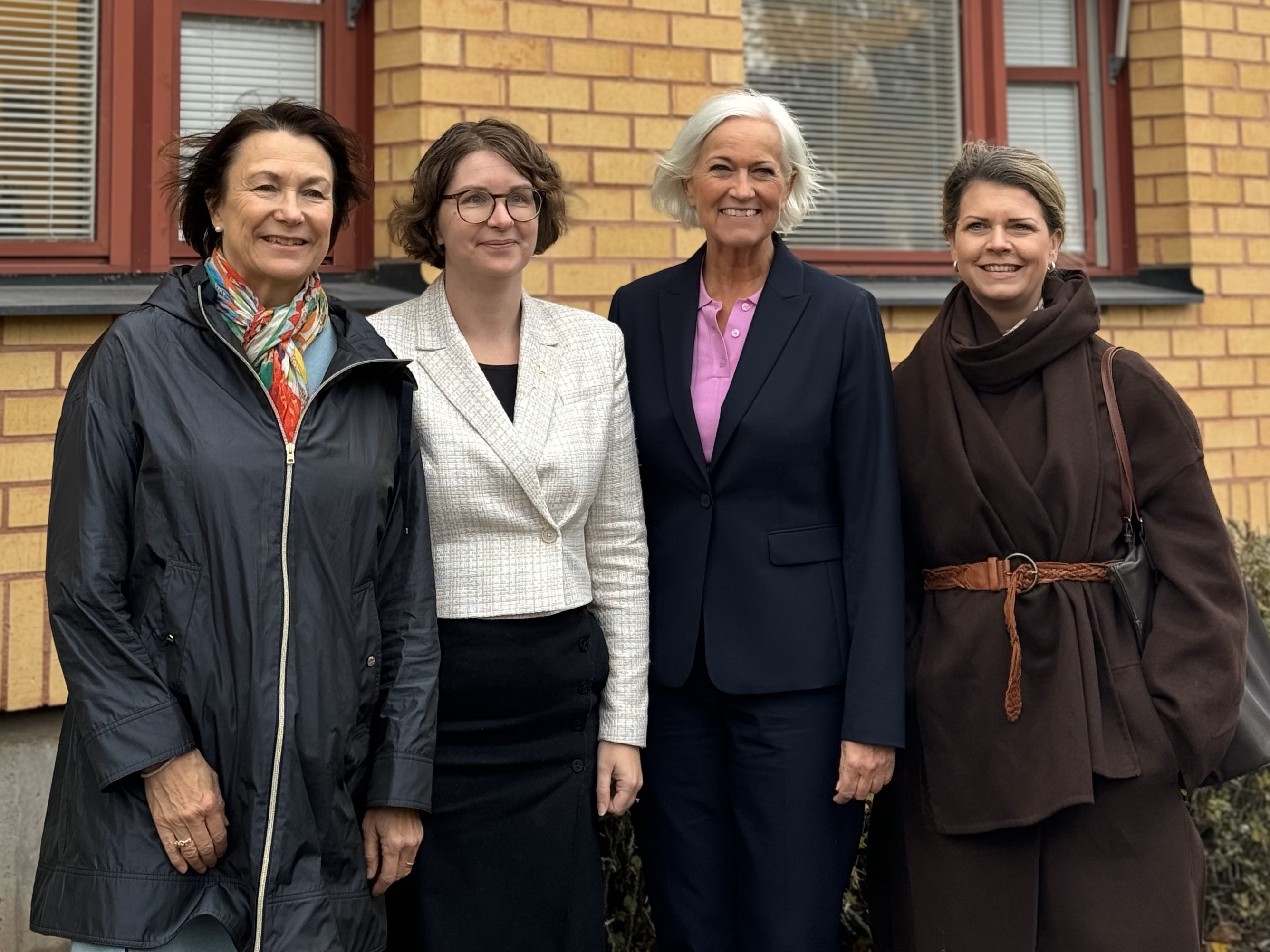 Fyra kvinnor står uppställda bredvid varandra framför en tegelvägg för att fotograferas. De är utomhus och har ytterkläder på sig.