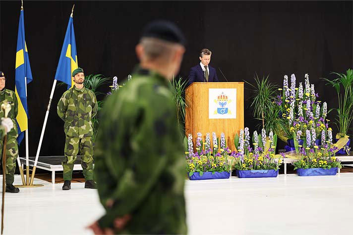 Till vänster står H.K.H. Prins Carl Philip i uniform framför två svenska flaggor. Centralt i bilden, ur fokus, är konturen av en man i uniform. Till höger står försvarsminister Pål Jonson i en talarstol och håller ett tal. Framför talarstolen står blommor.