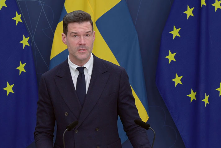 Migrationsminister Johan Forssell vid en talarpulpet med flaggor i fonden.