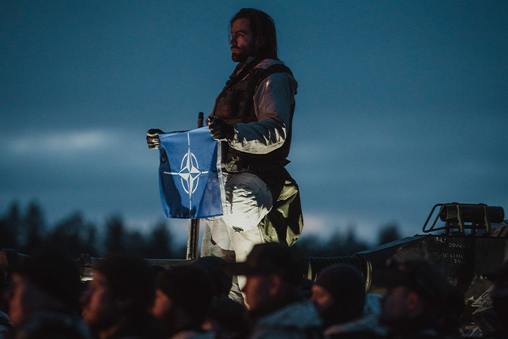 En samling soldater i kvällsljus. En soldat står upphöjt och håller i en Natoflagga.
