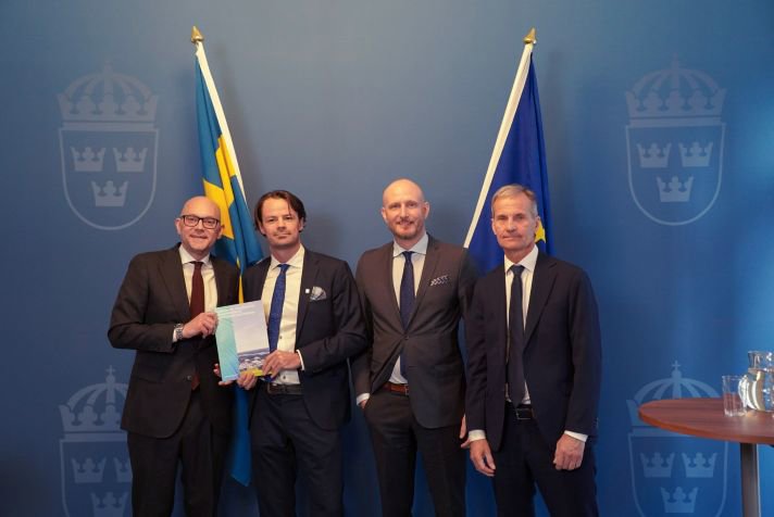 Ministern håller upp ansökan. De tre övriga står intill mot en bakgrund med RK:s logotyp.