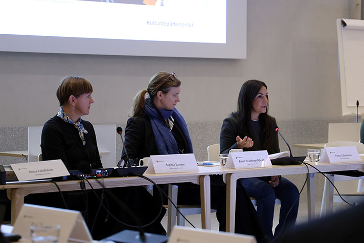 De tre personerna sitter på rad vid ett bord, kulturministern talar och övriga två tittar på henne.