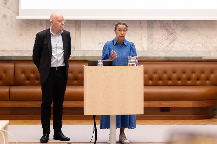 Meriam Chatty står vid en talarstol och pratar. Henrik Bachner står vid sidan om.