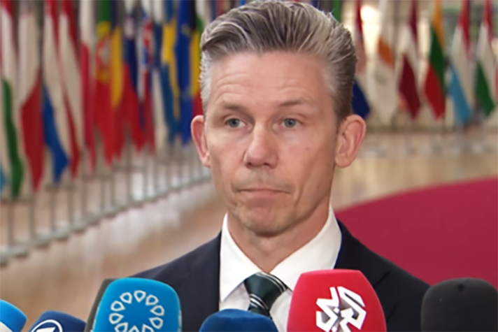 Pål Jonson står inomhus och pratar med media. I bakgrunden syns EU-ländernas flaggor.