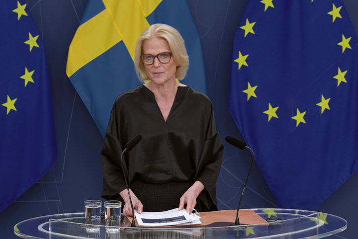 Statsrådet står vid ett ståbord med sveriges och EU:s fana i fonden