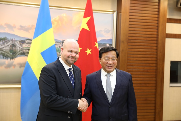 Två leende män i kostym skakar hand framför den svenska och kinesiska flaggan.