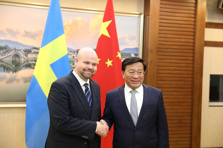 Två leende män i kostym skakar hand framför den svenska och kinesiska flaggan.