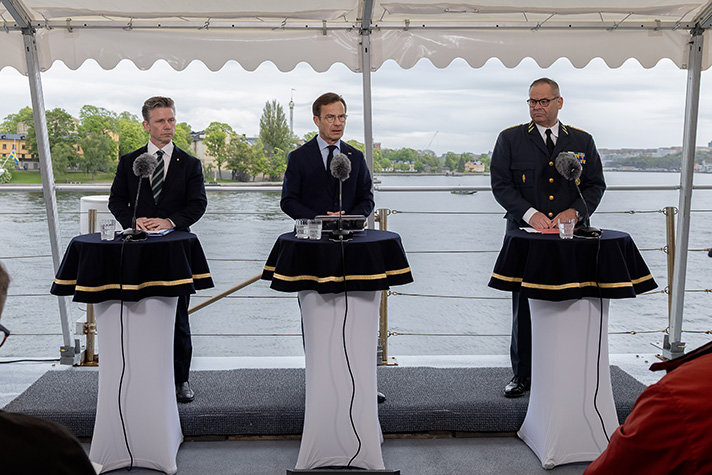 Försvarsminister Pål Jonson, statsminister Ulf Kristersson och överbefälhavare Michael Claesson står med ryggen till Strömmen och Skeppsholmen. Framför dem står ståbord prydd med marinblå duk. Ovanpå ståborden står mikrofoner.