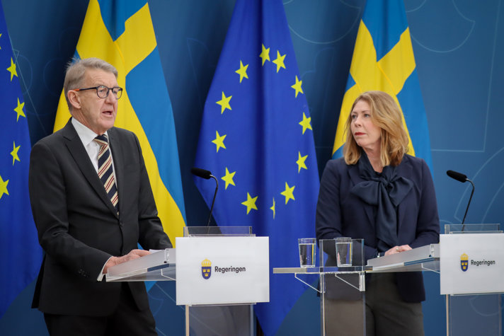 Anders Sylvan, samordnare för kortare vårdköer, står vid en talarpulpet och pratar. Blicken är riktad mot en publik vi inte ser. Bredvid honom står sjukvådsminister Elisabet Lann, vid en annan talarpulpet, och tittar på honom.