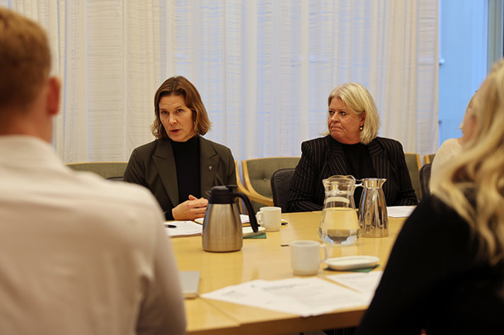 Jämställdhetsminister Nina Larsson och socialtjänstminister Camilla Waltersson Grönvall sitter vid ett bord. Nina talar och Camilla tittar på henne.