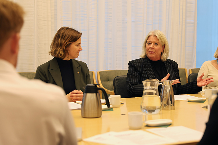 Jämställdhetsminister Nina Larsson och socialtjänstminister Camilla Waltersson Grönvall sitter vid ett konferensbord. Camilla talar och Nina tittar på henne.
