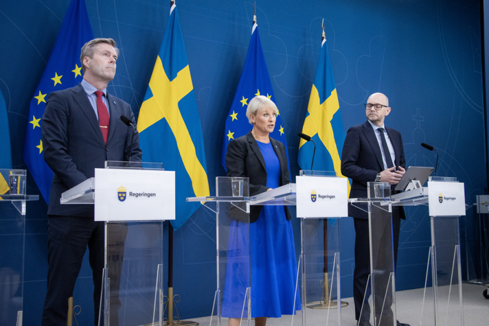 Anna Tenje, Johan Britz och Hans Eklind står vid varsin talarstol och framför svenska och EU-flaggan i regeringens pressrum.