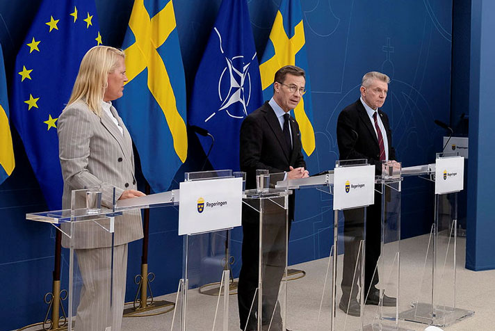 Tre personer, statsministern i mitten, fotograferade snett från vänster när de står vid var sin talarstol. Det är flaggor i fonden.