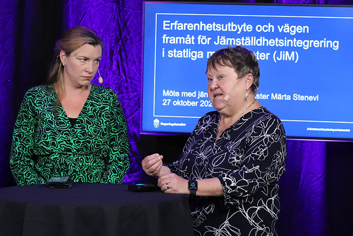 Ylva Strander står och kollar på Ingrid Petersson som talar. Bakom dem syns en skärm med texten Erfarenhetsutbyte och vägen framåt för Jämställdhetsintegrering i statliga myndigheter (JiM)