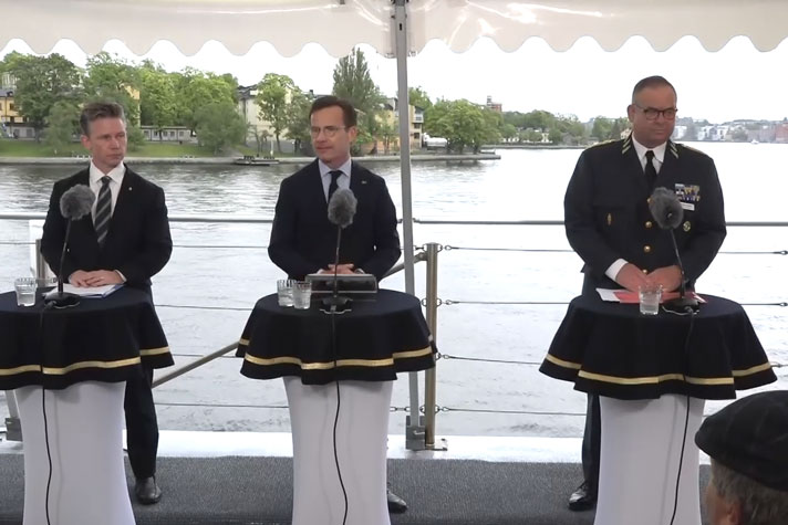 De medverkande står vid var sin talarpulpet på ett båtdäck. I bakgrunden ser man Skeppsholmen.