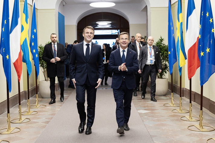 Emmanuel Macron och Ulf Kristersson går bredvid varandra inne i Rosenbad flankerade av svenska flaggor, franska flaggor och EU-flaggor. I bakgrunden syns fler personer.
