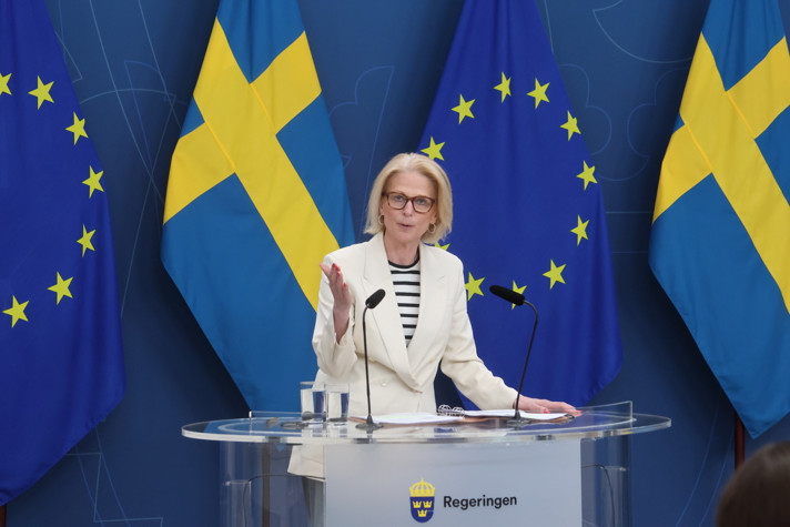 Finansministern talar inför journalister vid en pressträff. I bakgrunden syns Sverige- och EU-flaggor.