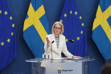 Finansministern talar inför journalister vid en pressträff. I bakgrunden syns Sverige- och EU-flaggor.