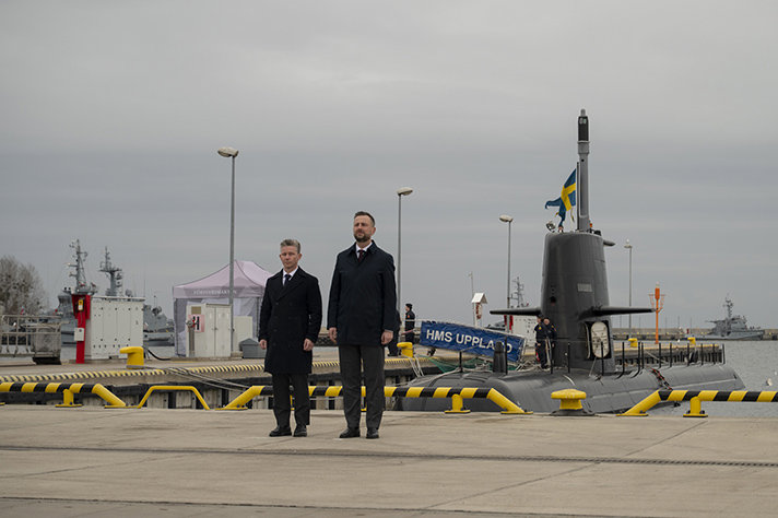 Pål Jonson och Władysław Kosiniak-Kamysz står på en kaj i Gdynia. Det är mulet och i bakgrunden syns en svensk ubåt ligga förtöjd.