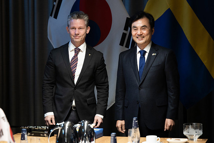 Pål Jonson och Ahn Gyu-back står sida vid sida vända mot kamerorna som inte syns i bild. I bakgrunden syns Sydkoreas och den svenska flaggan.