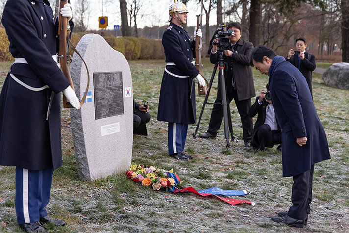Ahn Gyu-back bugar framför en minnessten. Framför minnesstenen ligger en krans. Minnesstenen pryds med ett plakat och den svenska samt den sydkoreanska flaggan. På varsin sida av stenen står svenska hedersvakter i givakt. I bakgrunden syns också koreanska fotografer.