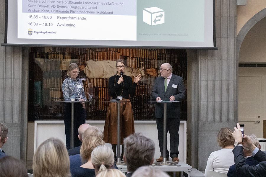 Tre personer står på en scen och pratar framför en publik