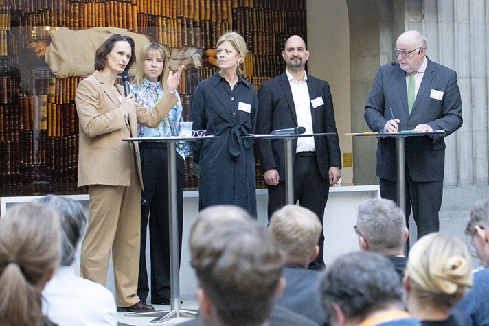 Fem personer står på en scen och pratar framför en publik