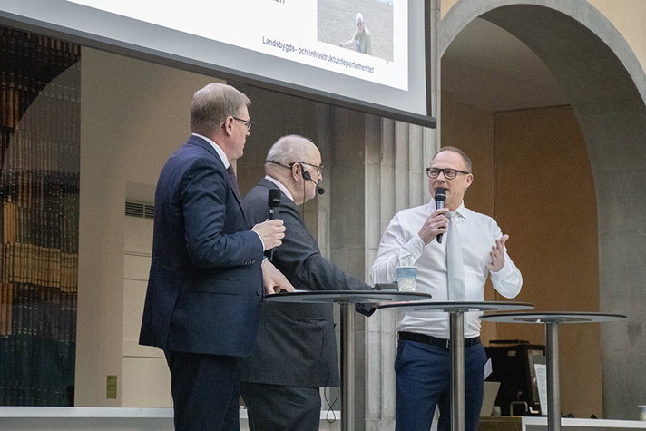 Tre personer står på en scen och pratar framför en publik