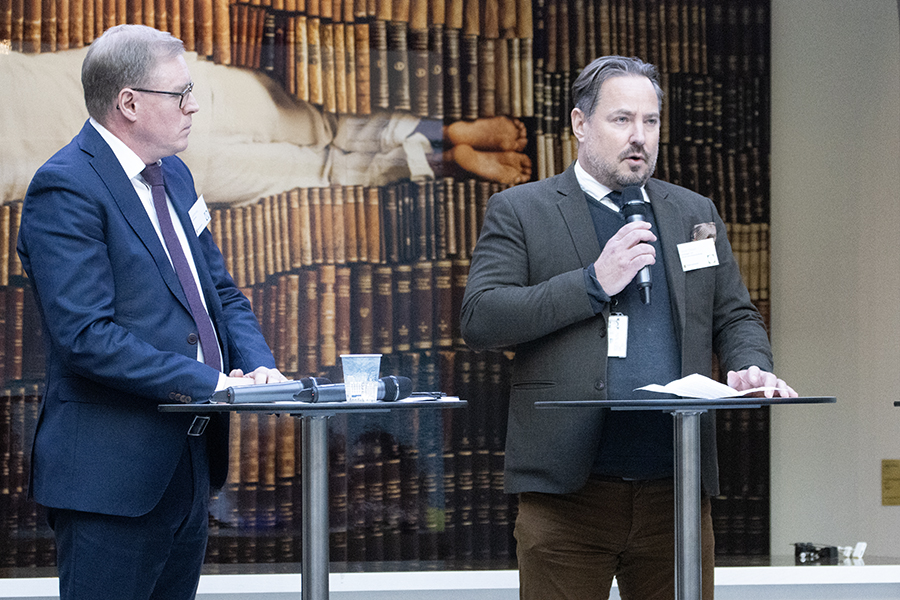 Två personer står på en scen och pratar framför en publik