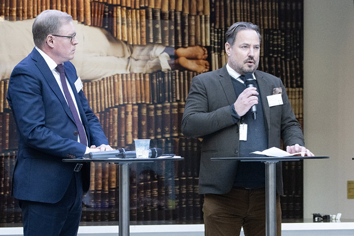 Två personer står på en scen och pratar framför en publik