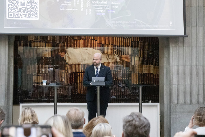 En man i mörk kostym står på en scen och pratar framför en publik