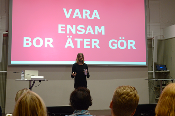 Micael Dahlen föreläser inför publik. Bakom honom finns en stor skärm som visar orden "Vara ensam. Bor. Äter. Gör."