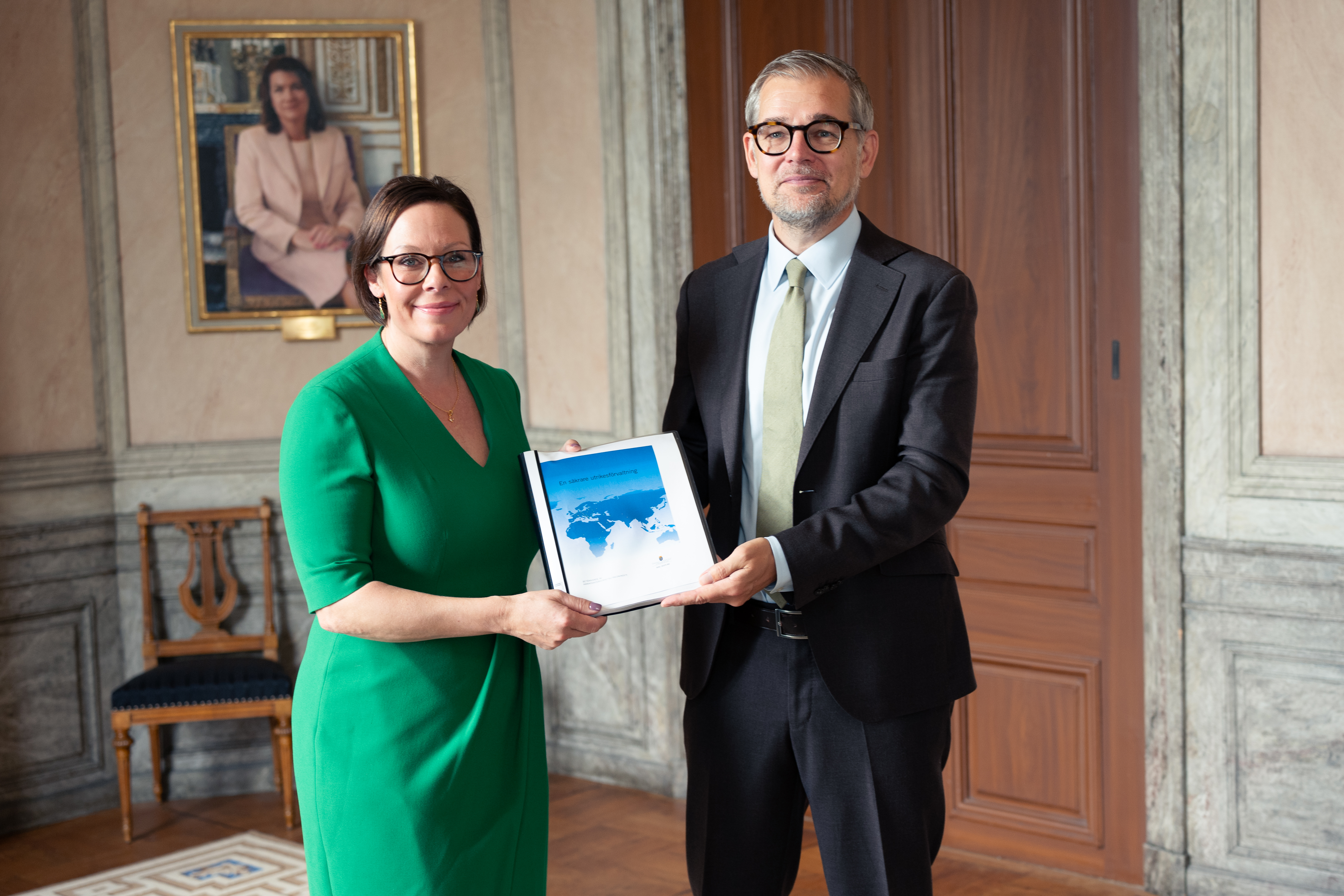 Kvinna i grön klänning och man i svart kostym hållet upp en bunt papper mellan sig.