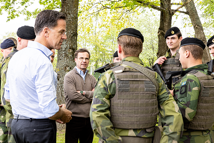 Mark Rutte och Ulf Kristersson står i naturen och talar med militärer.