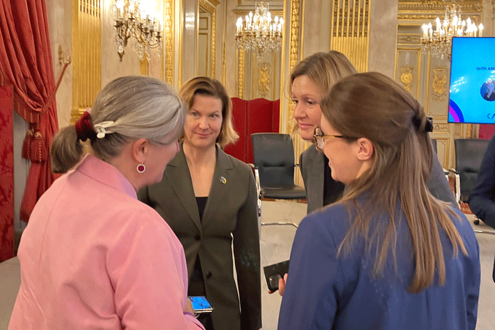 Nina Larsson samtalar med tre kvinnor. I bakgrunden syns ett förgyllt rum med ljuskronor och röda sammetsgardiner.