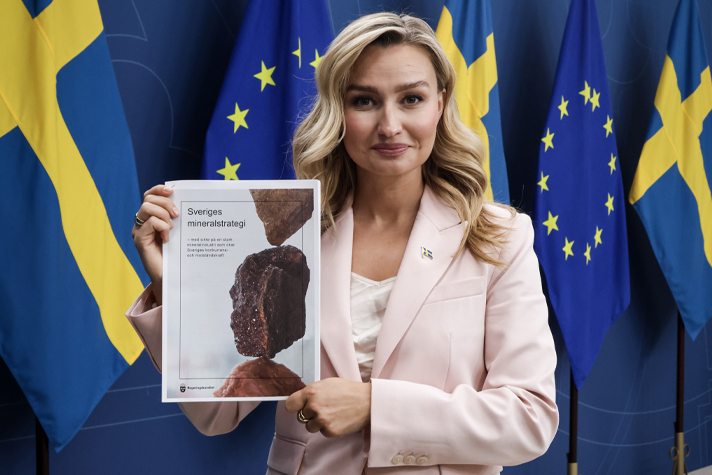 Energi- och näringsminister Ebba Busch håller upp Sveriges mineralstrategi i tryckt form. Busch är klädd i rosa kavaj och står fram för en blå bakgrund med flaggor.