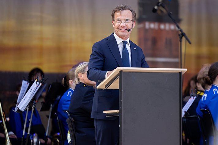 Ulf Kristersson står i en talarstol på en utomhusscen. I bakgrunden syns en orkester.