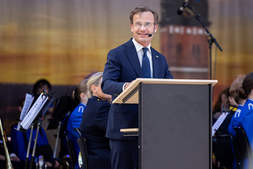Ulf Kristersson står i en talarstol på en utomhusscen. I bakgrunden syns en orkester.