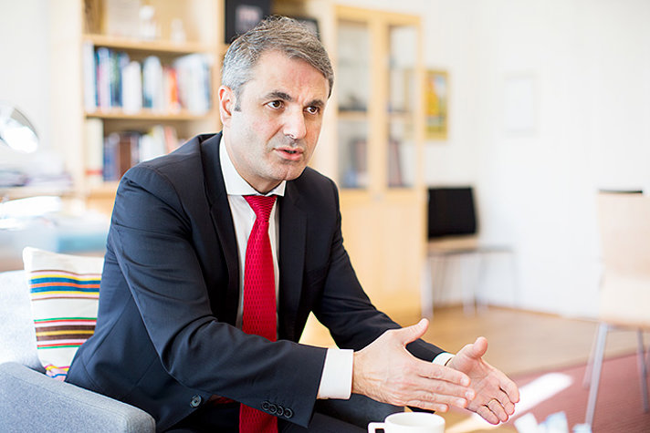 Ibrahim Baylan sitter i en fåtölj. Han är klädd i blå kostym, vit skjorta och röd slips.