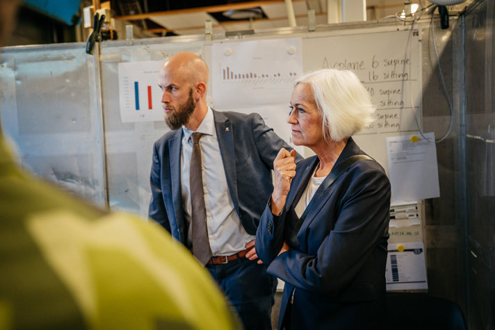 Två personer står i ett rum och lyssnar på en person som pratar.