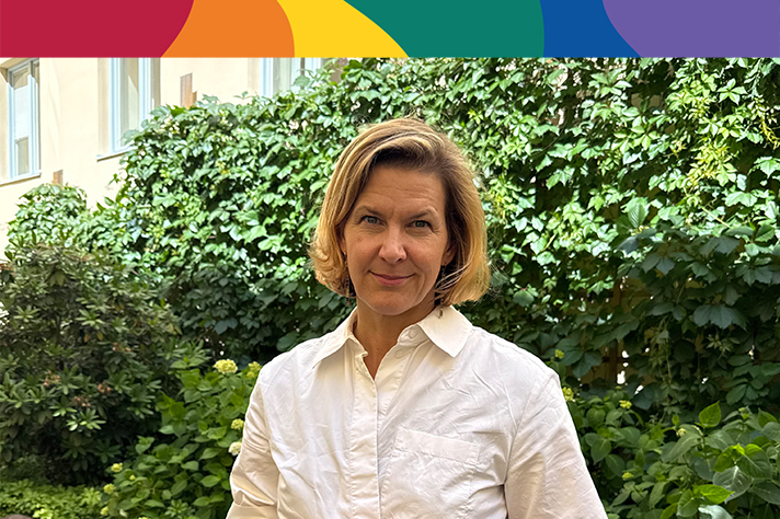 En kvinna som står framför gröna buskar. Ovanför fotot finns en banner i regnbågens färger.