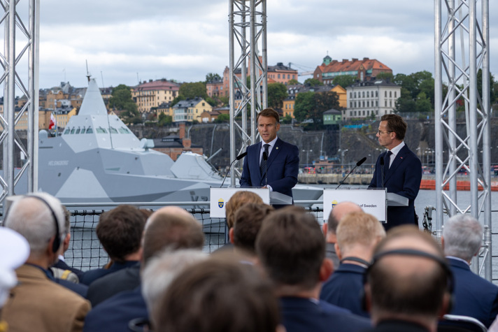 Emmanuel Macron och Ulf Kristersson står vid varsin talarstol ombord på en fregatt. I bakgrunden syns en militär båt samt Södermalms siluett.