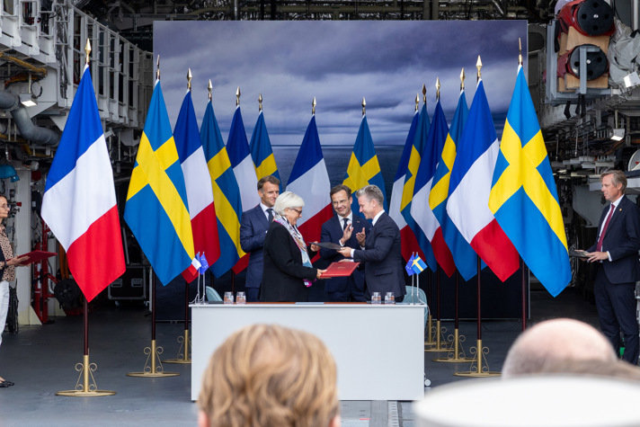 Emmanuel Macron, Ulf Kristersson, Catherine Vautrin och Pål Jonson vid ett signeringsbord. I bakgrunden svenska och franska flaggor.