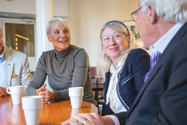 Inomhusbild där Tenje sitter vid runt bord och samtalar med två äldre personer. På bordet står vita kaffekoppar.