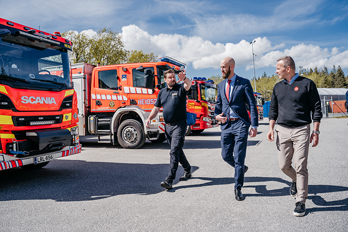 Utomhusbild. Bohlin flankerad av två personer går aktivt snett mot kameran. Till vänster i bild står räddningsfordon uppställda. Blå himmel med vita tussiga moln.